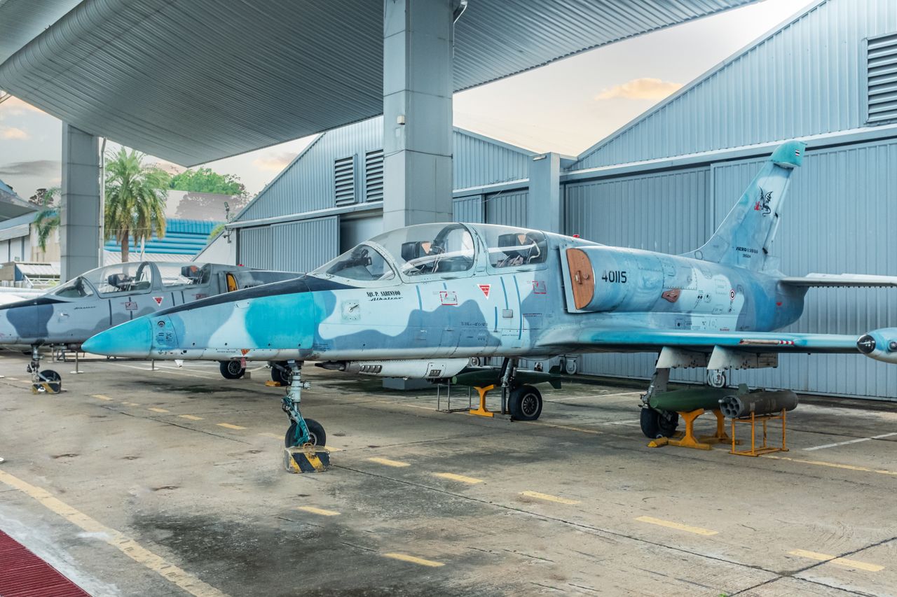 Two military training jets parked under a metal shelter at an airbase, prepared for maintenance.