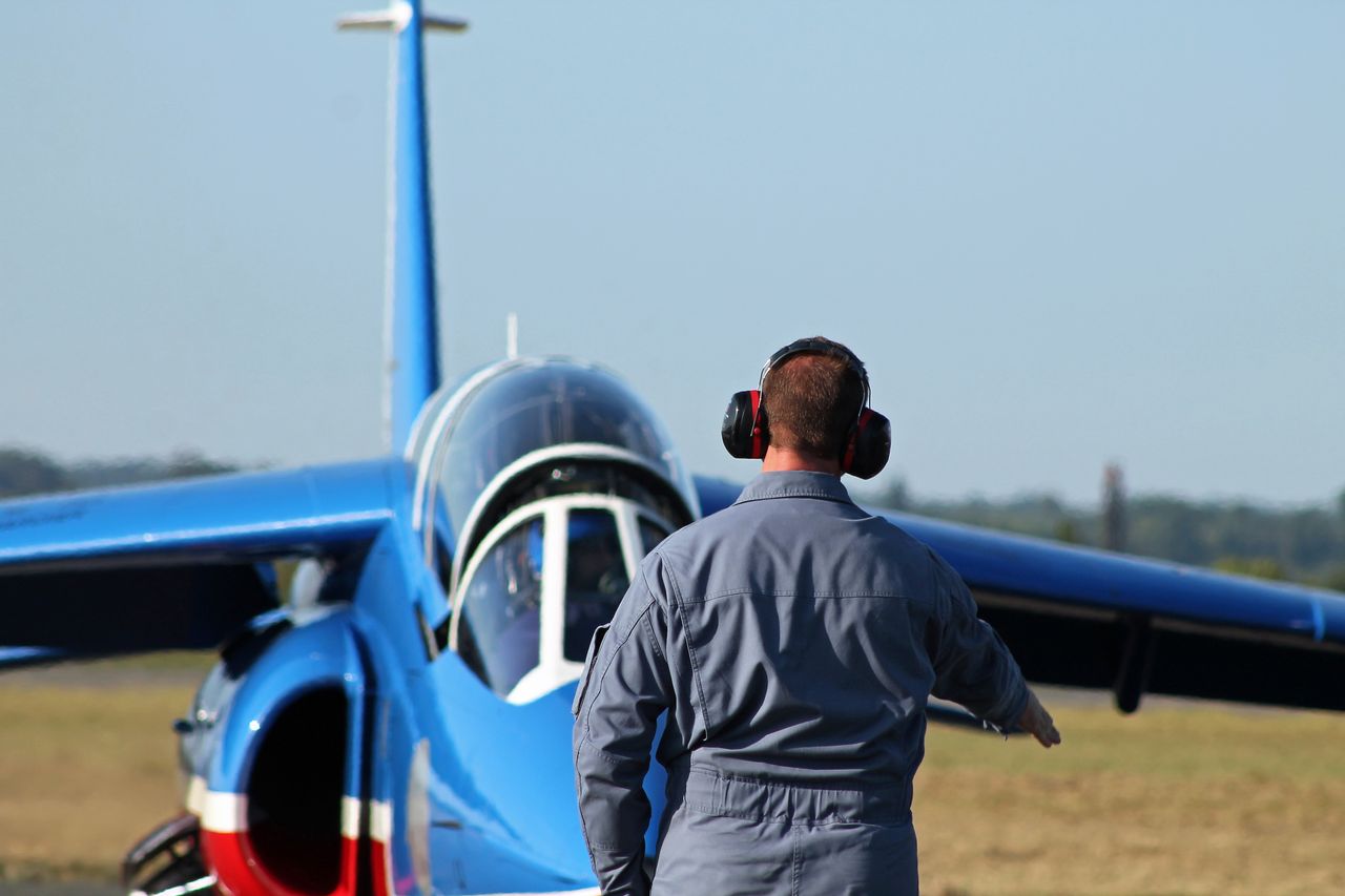 Aircraft technician guides blue jet on runway with headphones for noise reduction.
